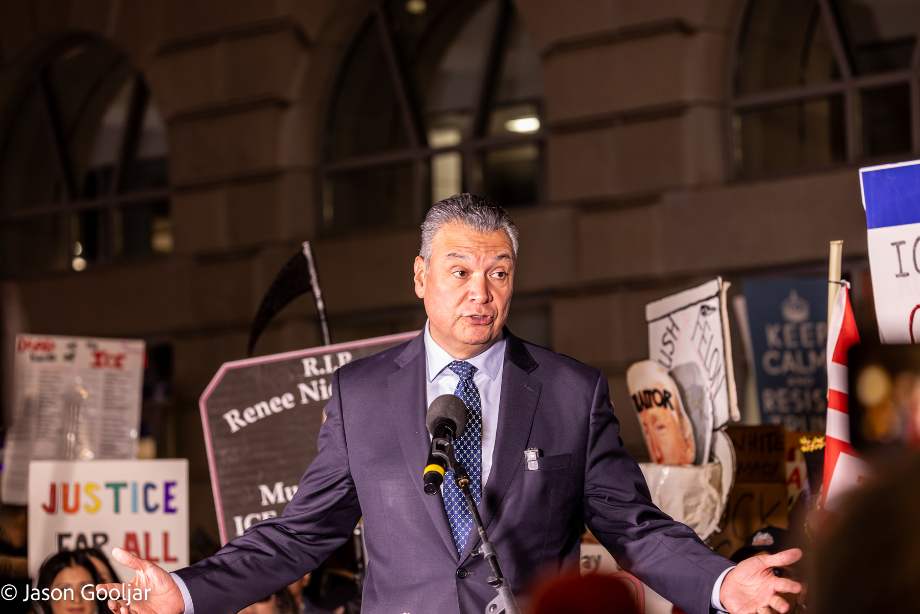 A man in a suit stands at a microphone, surrounded by numerous signs and placards at a protest or rally.