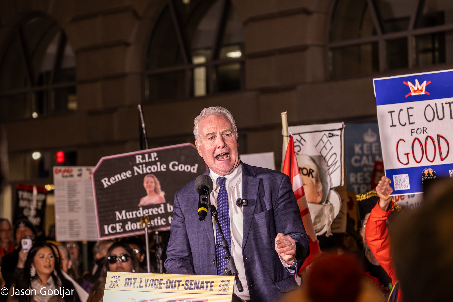 A person is speaking at a rally or demonstration with people holding signs in the background.