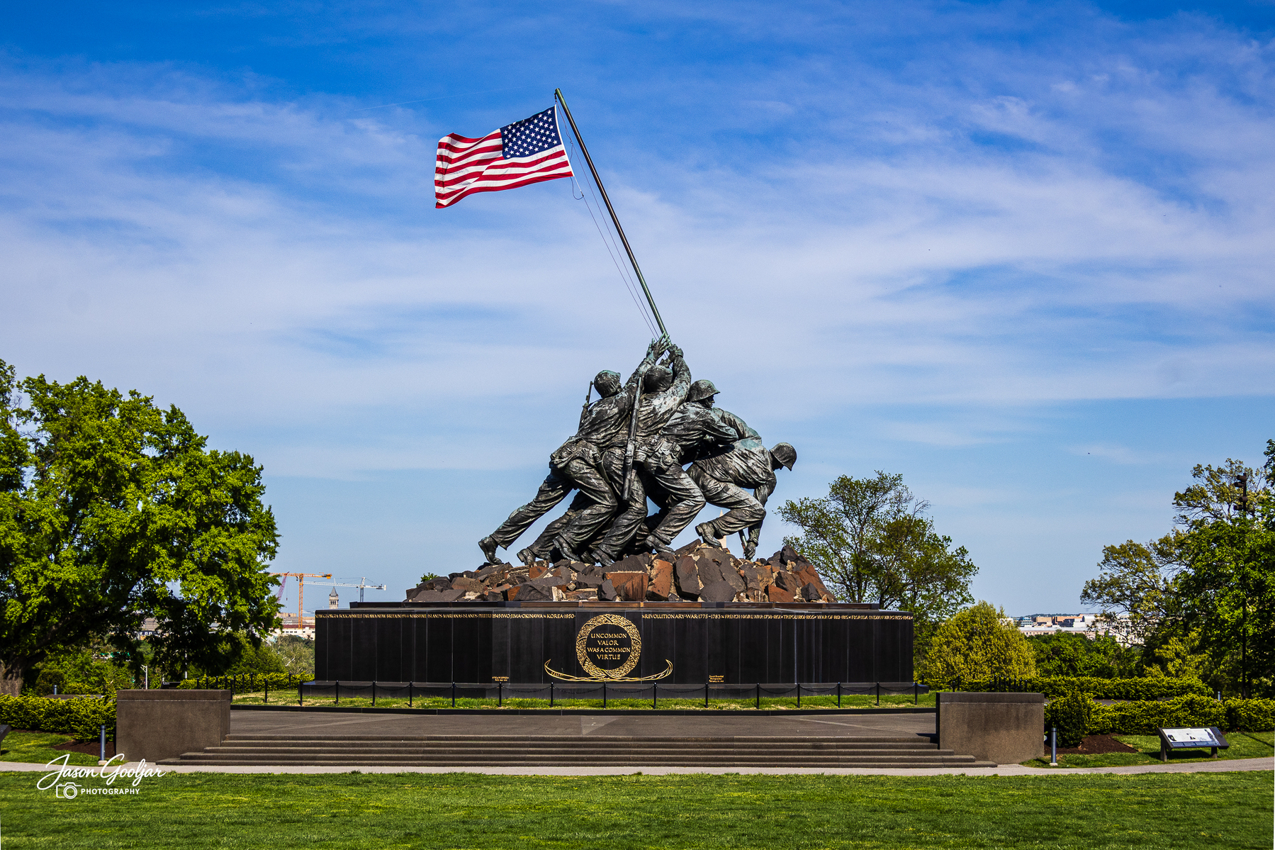 Several soldiers are depicted raising an American flag in a monument surrounded by greenery and a clear blue sky.
